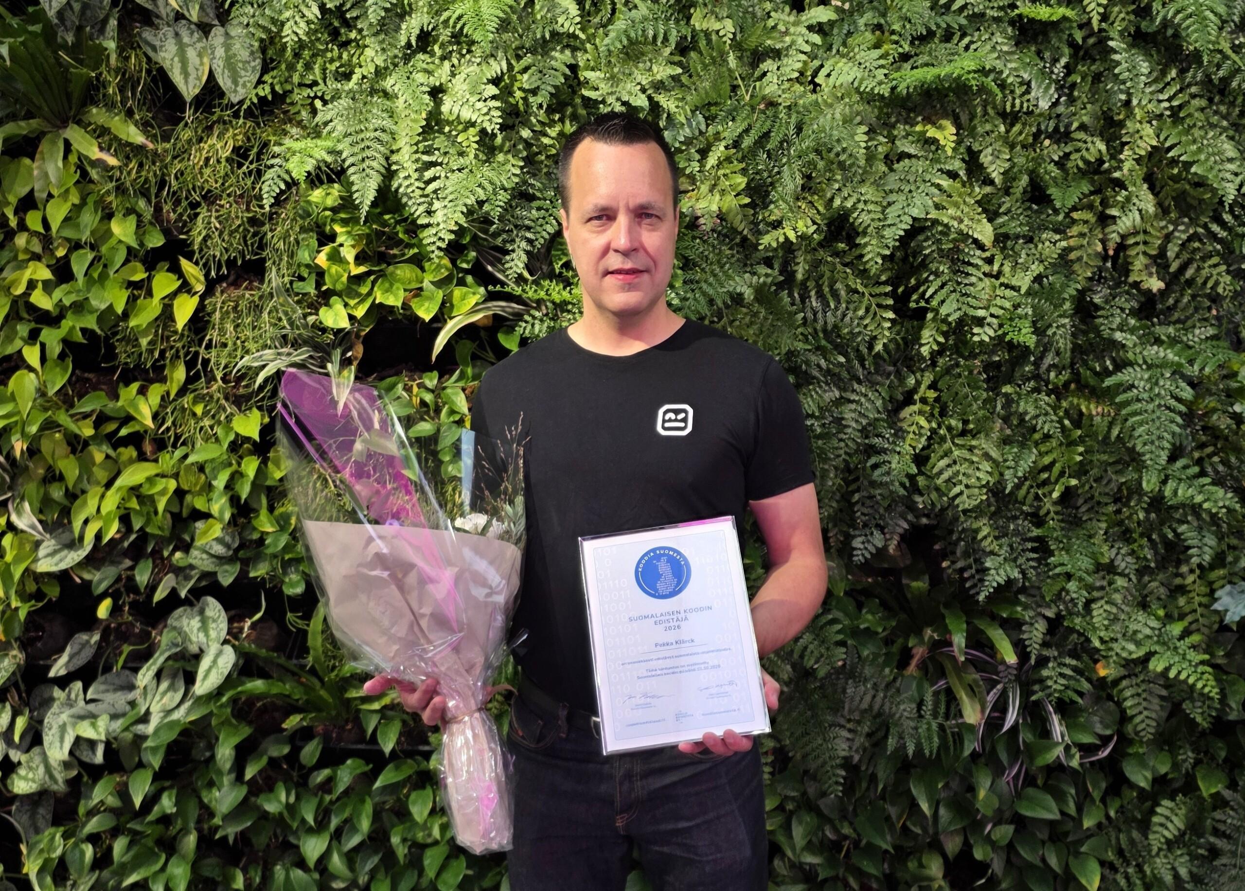 Pekka Klärck pitelee käsissään Suomalaisen koodin edistäjä -diplomia ja kukkakimppua.